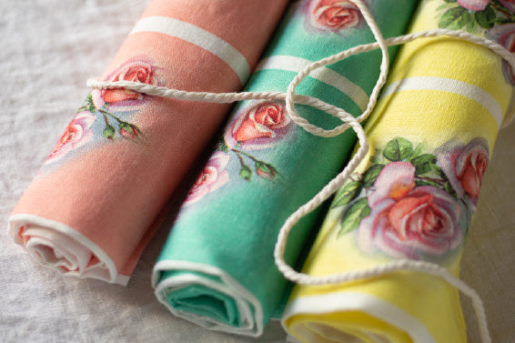 Showing a set of 3 rolled up linen tea towels with roses on them and three different coloured backgrounds which make a lovely set. Colours are a pastel blue/green aqua, a bright lemon yellow and a dusky salmon pink. All with bright roses on them.