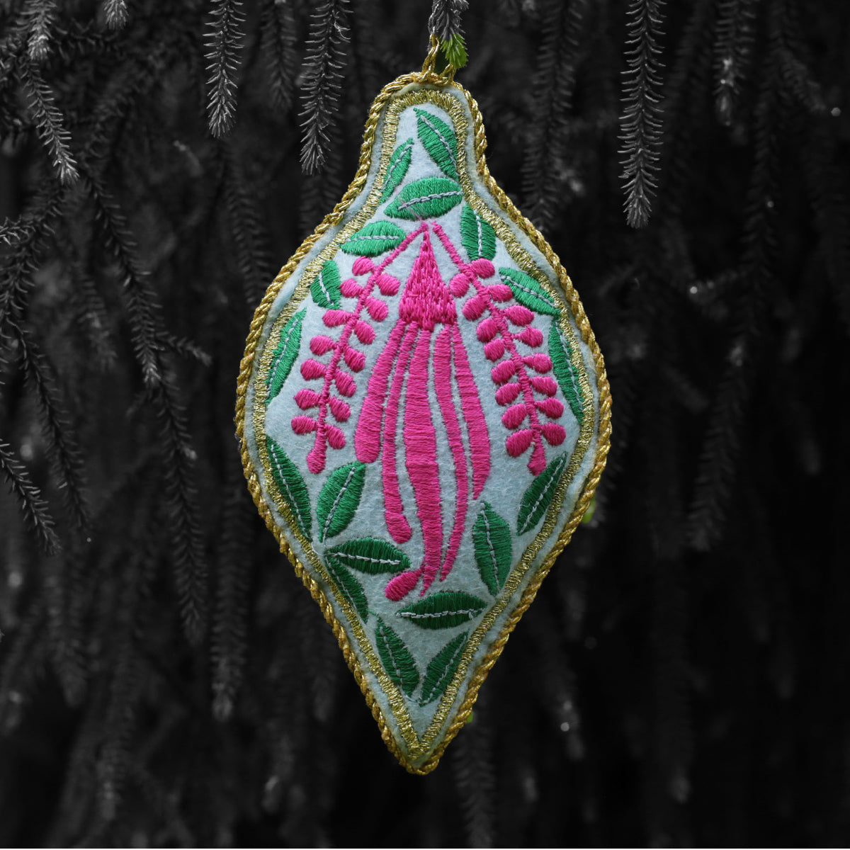 A red and green embroidered Christmas tree ornament with leaves and pinnk flower, hanging from a branch.