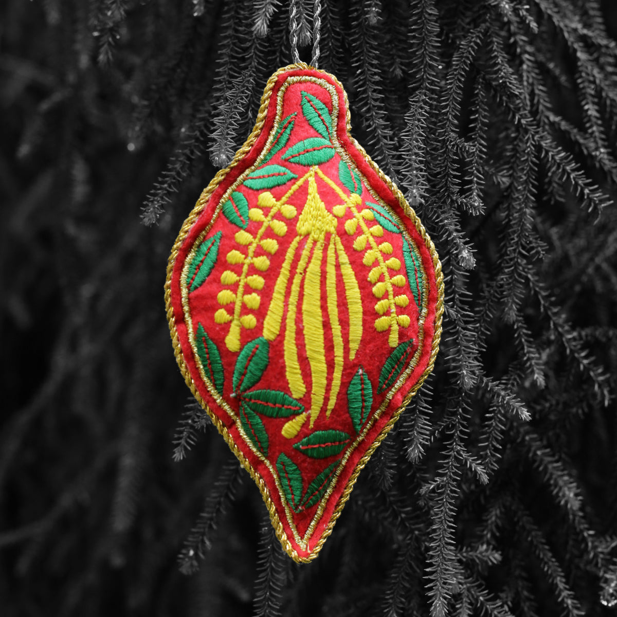 A red and green embroidered Christmas tree ornament with leaves and yellow flower, hanging from a branch.