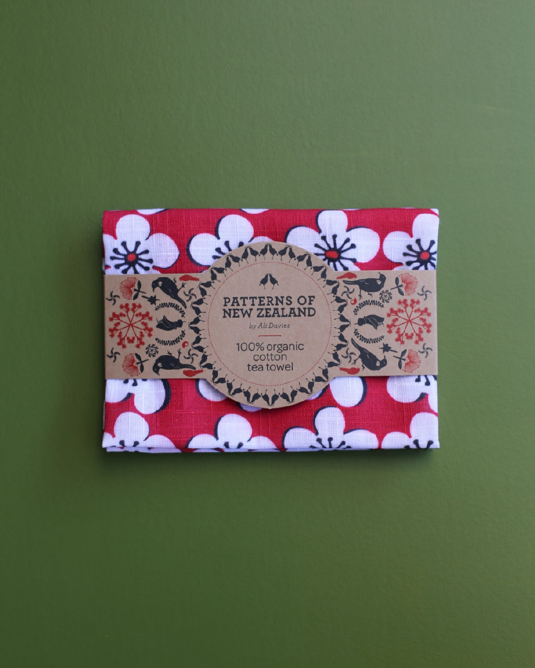 A folded cotton tea towel with a red, white, and black floral pattern, wrapped with a brown paper band labelled 'Patterns of New Zealand'. The towel is displayed flat against a solid olive green background.