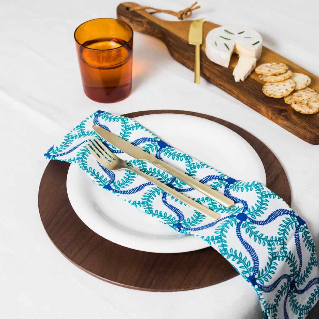 A set of two blue floral patterned cloth napkins arranged on a white plate with a fork and knife, displayed on a wooden table with a cheese platter in the background.