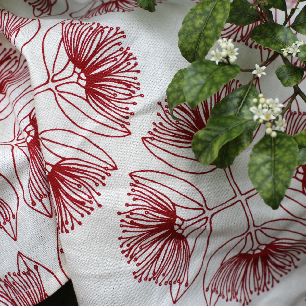Closeup of pohutukawa print in red fabric on a natural base with leaves of a trees resting on the fabric