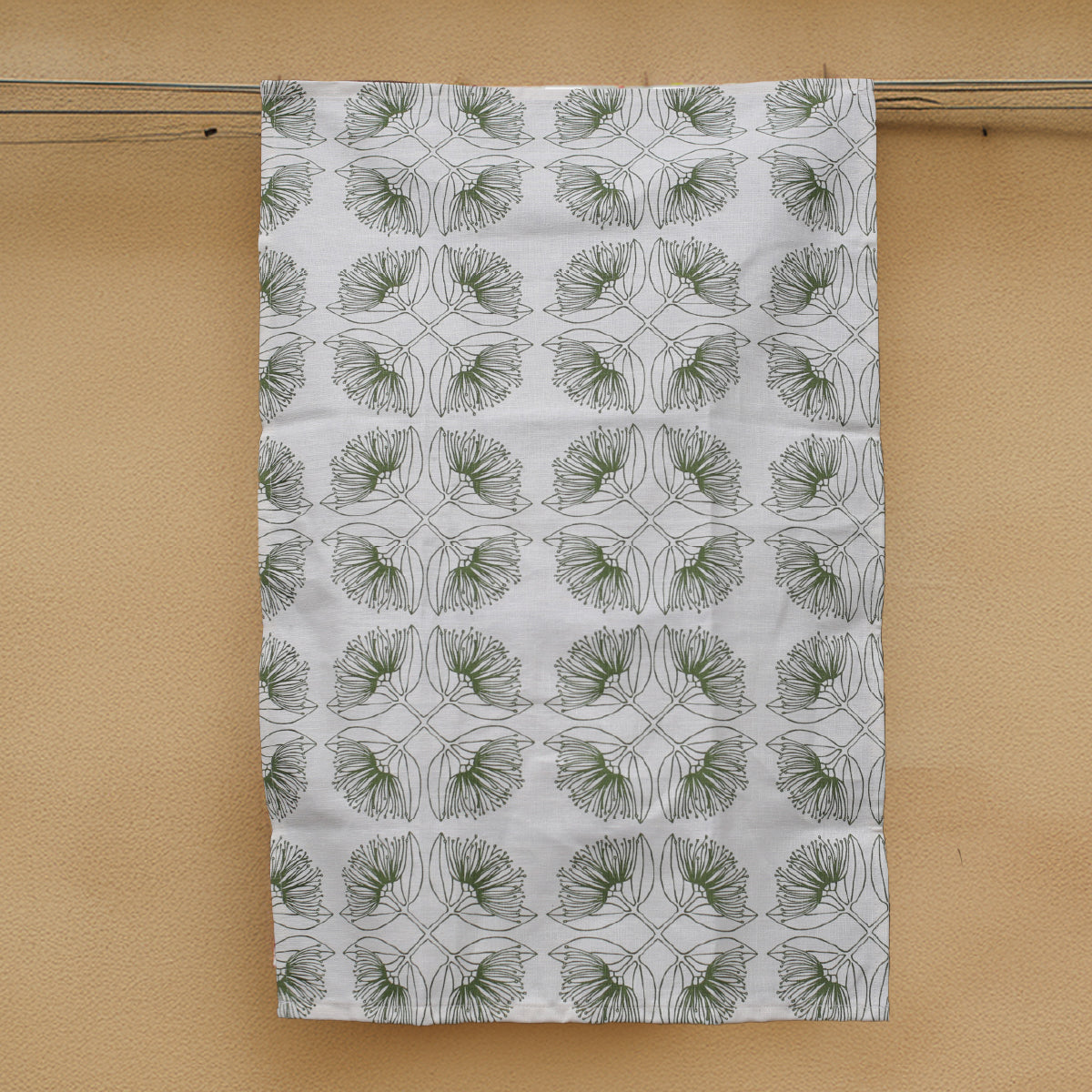 A white hemp tea towel with an olive green Pohutukawa flower print, hanging on a line against a beige wall.