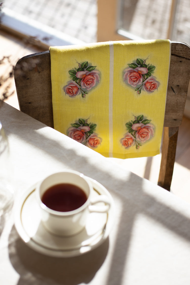 A vintage style yellow linen tea towel with a pink and green rose motif repeated vertically in four stripes, is folded over a wooden chair with a tea cup on the table.