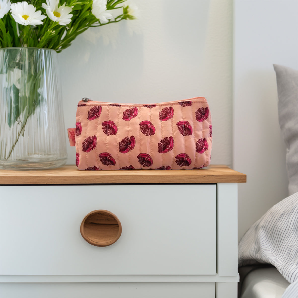 Small pink floral toilet bag sitting on a bedside table next to a vase of flowers.