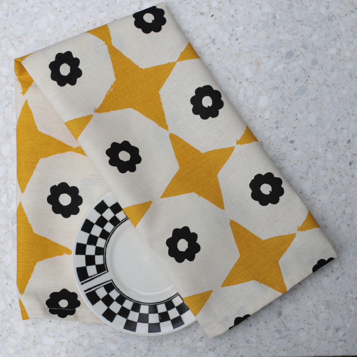 Closeup of folded yellow star and black flower printed hemp tea towel on a marble benchtop with black & white plate.