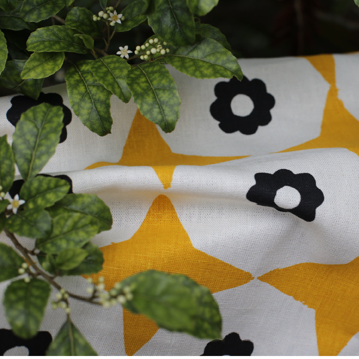 Closeup of yellow star and black flower printed fabric on a natural base with leaves of a trees resting on the fabric