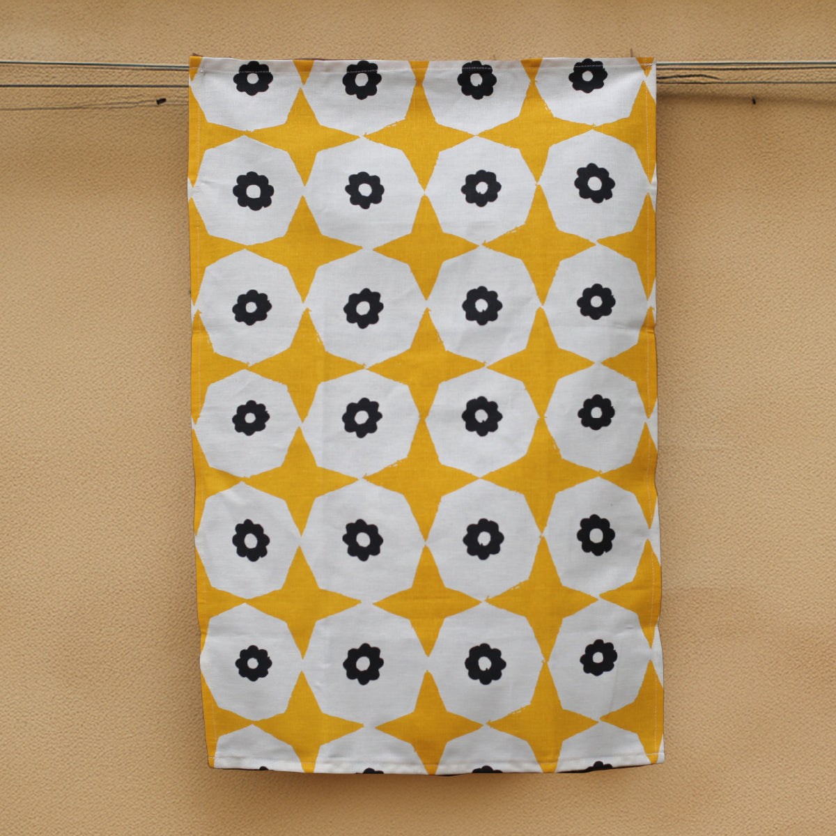 A yellow star and black flower printed hemp tea towel handing next to a honey coloured concrete wall.