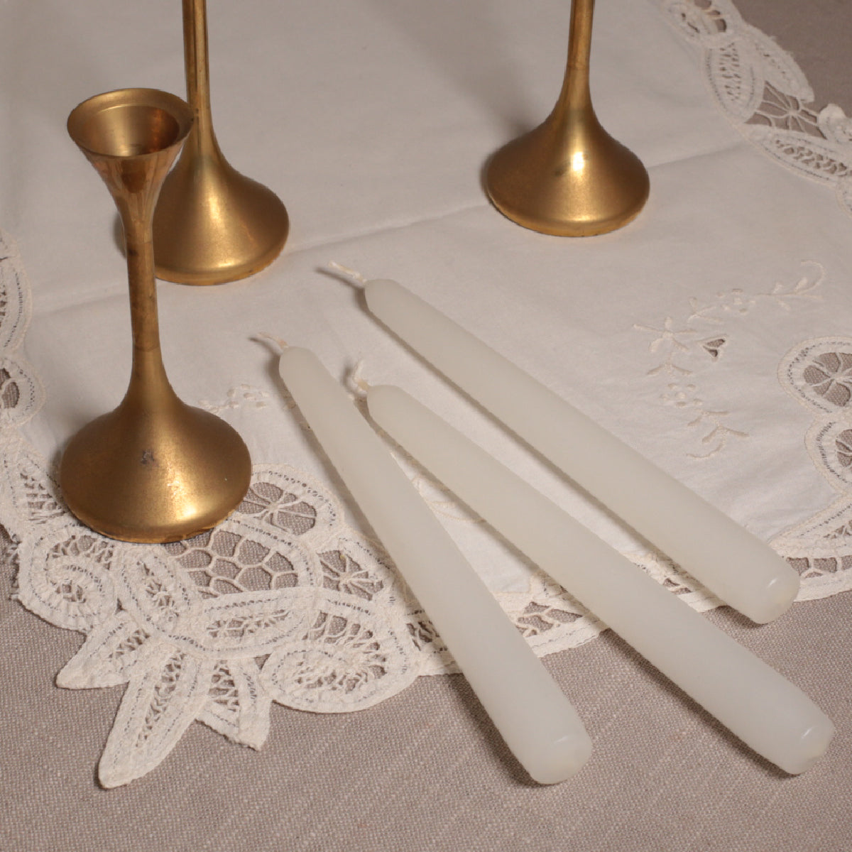 A set of three white Venetian-style tapered candles with gold-colored candleholders displayed on a lace tablecloth.