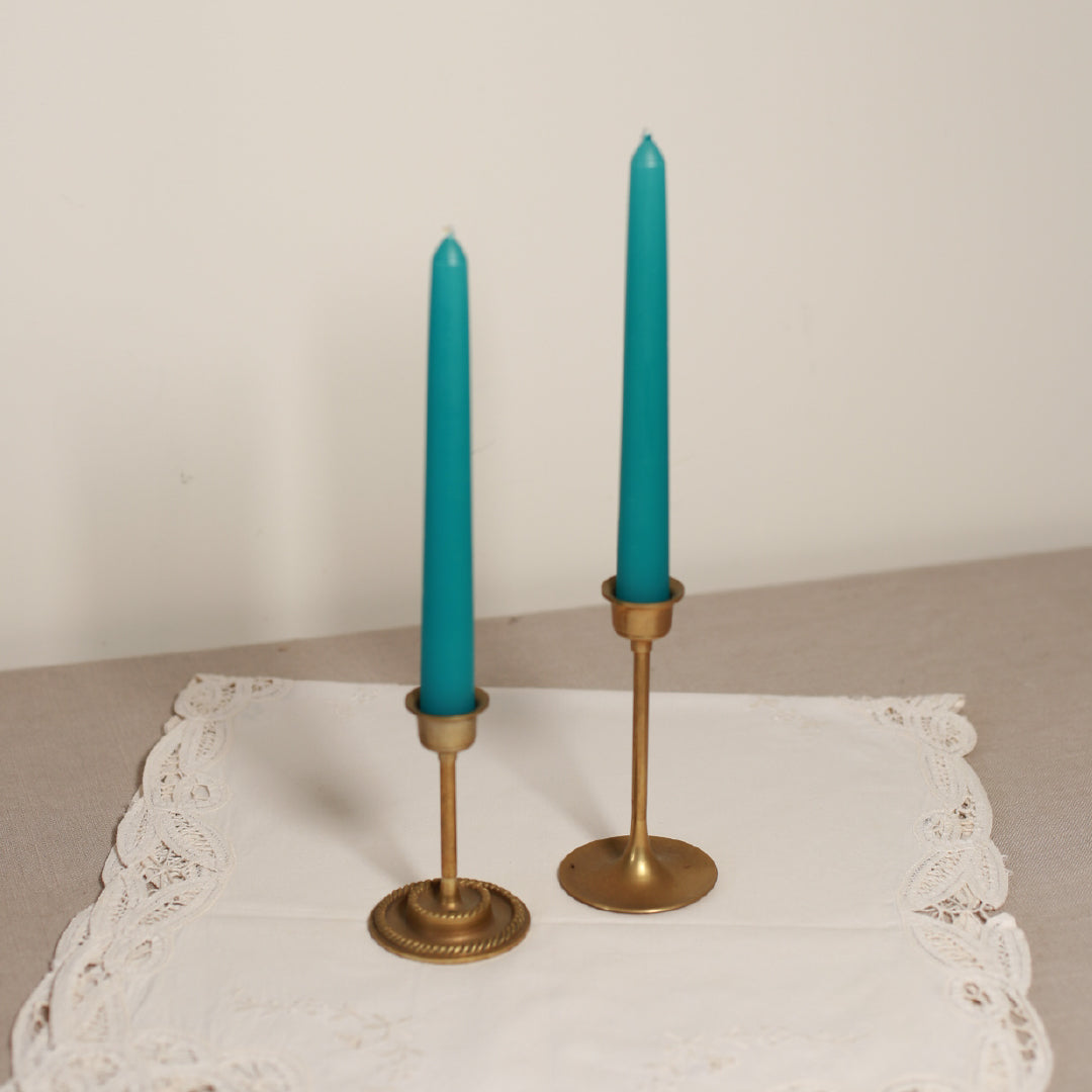 Two tapered Venetian-style table candles with turquoise wicks and bronze metal bases, placed on a lace-covered surface.
