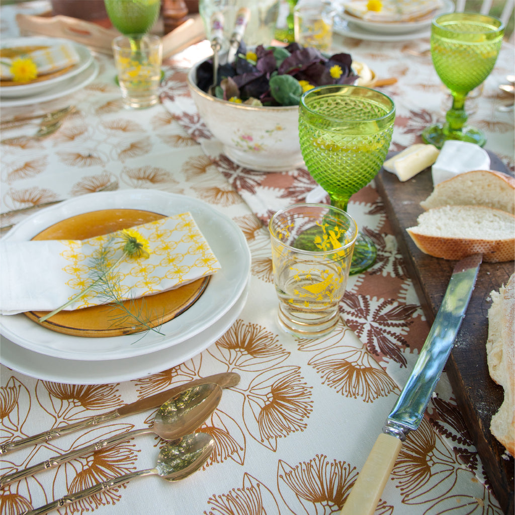 A closeup of a table setting with a pohutukawa patterned tablecloth in a dusk, sandy colour with green glasses, salad and bread and yellow printed napkins with a flower on them.
