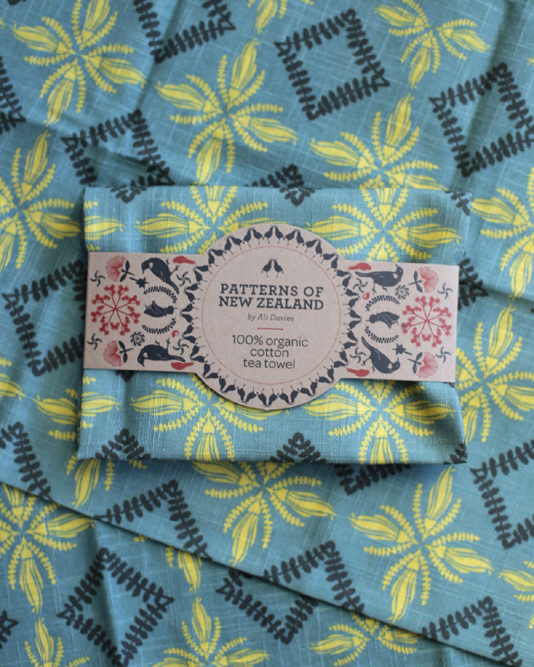 A folded cotton tea towel with a blue, yellow, and black geometric floral pattern, wrapped with a paper label that reads 'PATTERNS OF NEW ZEALAND by Misi Designs 100% organic cotton tea towel'.