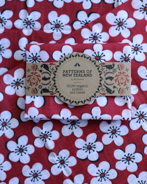 A folded cotton tea towel with a red, white, and black floral pattern, wrapped in a paper band labeled 'PATTERNS OF NEW ZEALAND 100% organic cotton tea towel'.