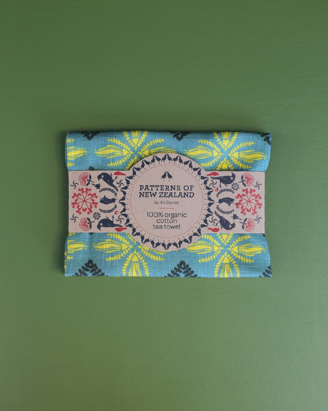 A folded cotton tea towel with a colorful floral and geometric pattern, wrapped in a paper band that reads 'PATTERNS OF NEW ZEALAND' and '100% organic cotton tea towel', set against a solid green background.