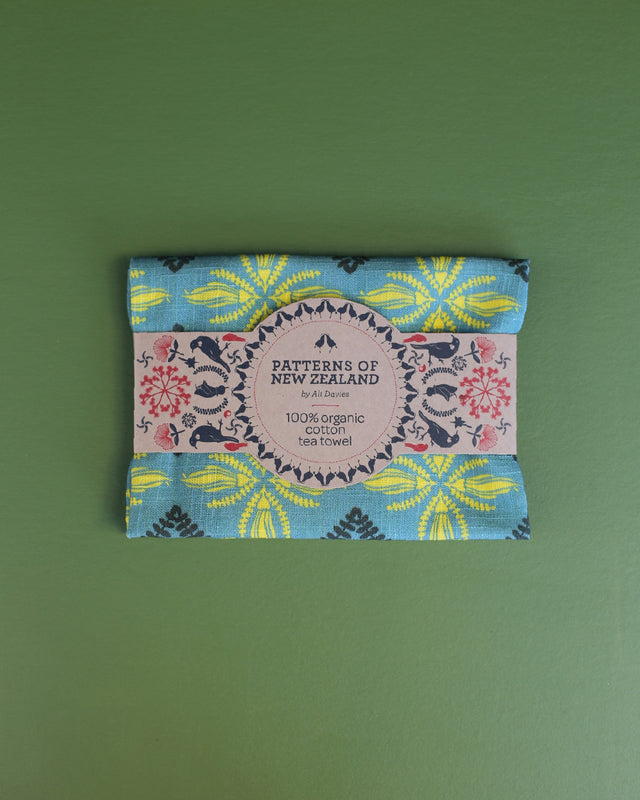 A folded cotton tea towel with a colorful floral and geometric pattern, wrapped in a paper band that reads 'PATTERNS OF NEW ZEALAND' and '100% organic cotton tea towel', set against a solid green background.
