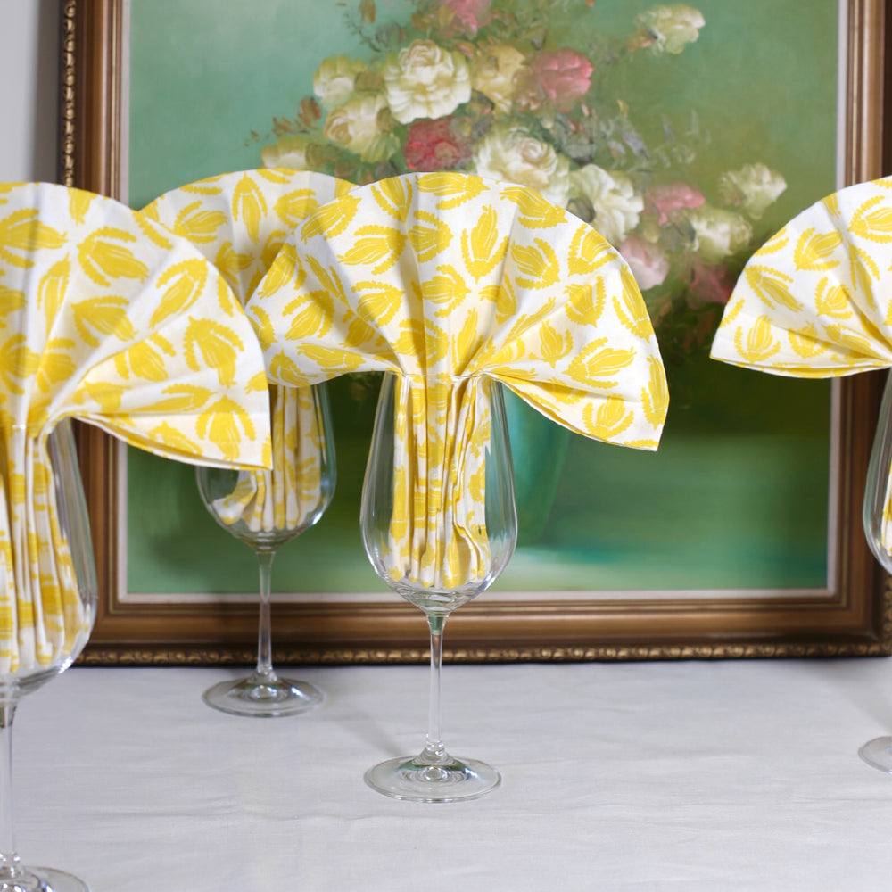 Four wine glasses with yellow napkins in a fan shape coming out of the glasses. There is a green floral still life painting behind.