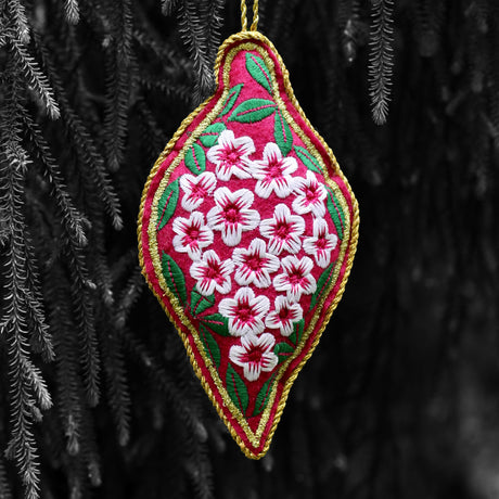 A triangular embroidered ornament with a pink base and a white floral pattern, hanging from a tree.