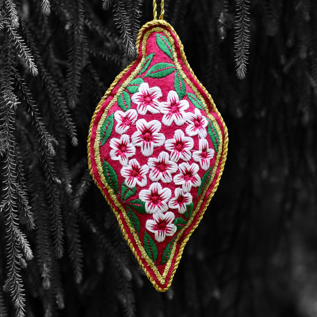 A triangular embroidered ornament with a pink base and a white floral pattern, hanging from a tree.