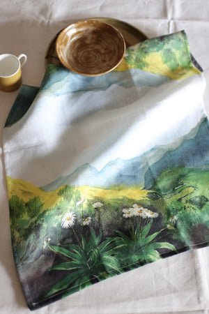 A tablecloth with a vibrant floral and mountainous landscape design, laid out on a table with a bowl and a cup.