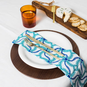 A set of two blue floral patterned cloth napkins arranged on a white plate with a fork and knife, displayed on a wooden table with a cheese platter in the background.