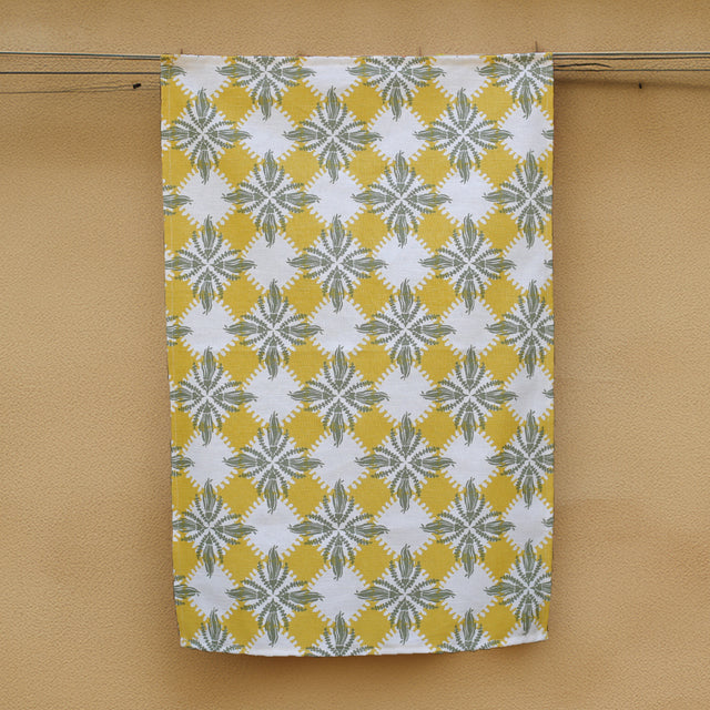 A hemp tea towel with a geometric Pacifica Kowhai pattern in olive green and Ceylon yellow, hanging on a yellow wall.