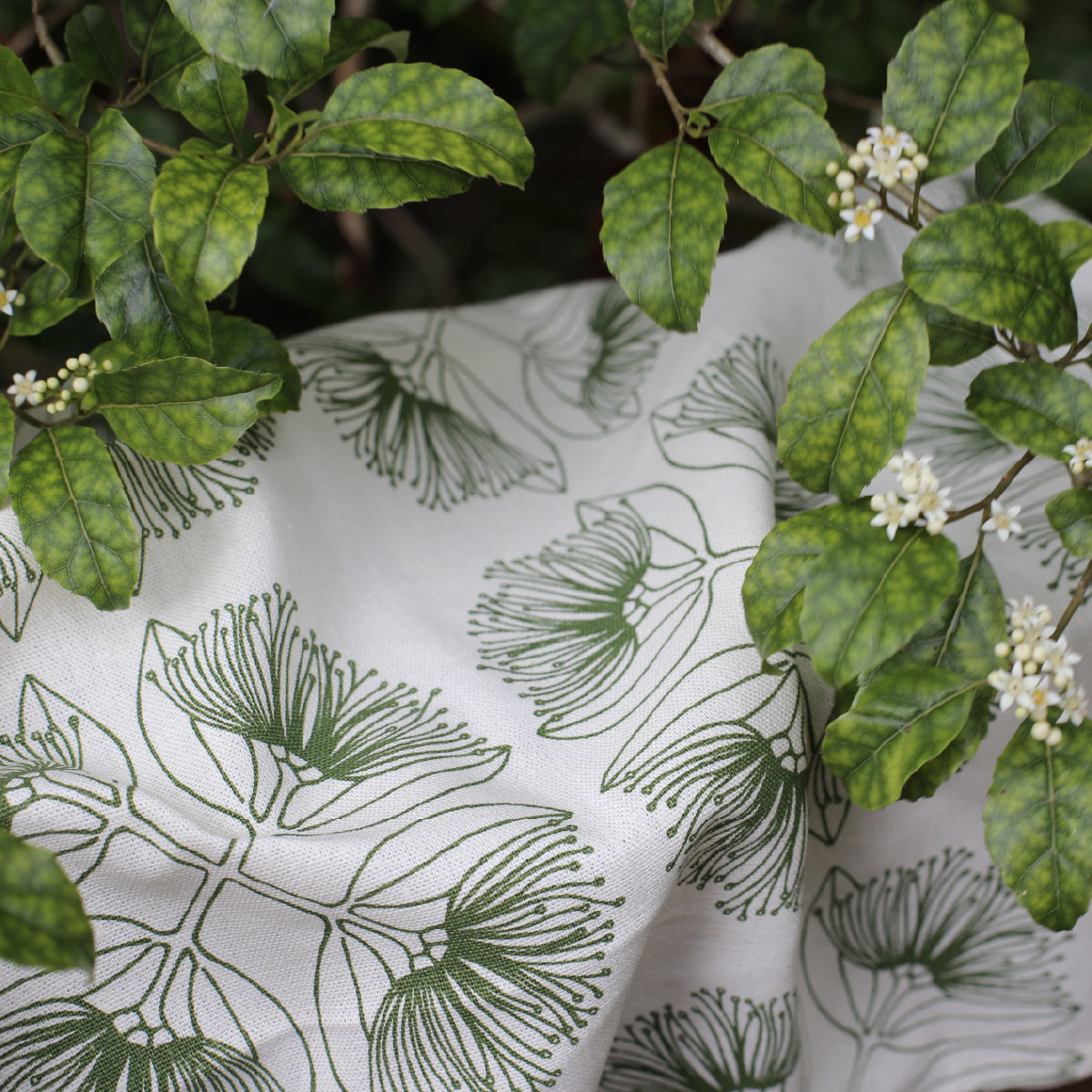 Closeup of pohutukawa print in olive  fabric on a natural base with leaves of a trees resting on the fabric