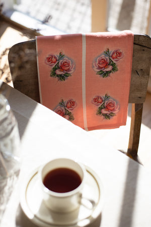A vintage style pink linen tea towel with a pink and green rose motif repeated vertically in four stripes, is folded over a wooden chair with a tea cup on the table.