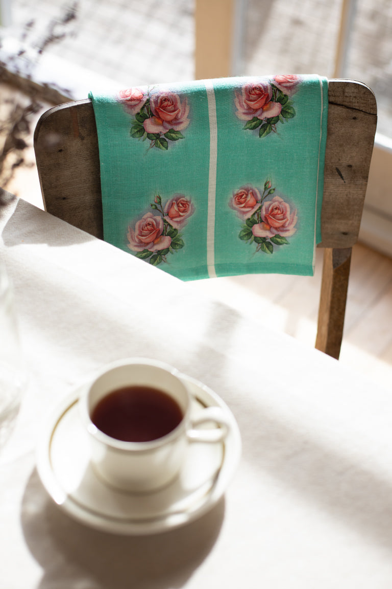 A vintage style teal linen tea towel with a pink and green rose motif repeated vertically in four stripes, is folded over a wooden chair with a tea cup on the table.