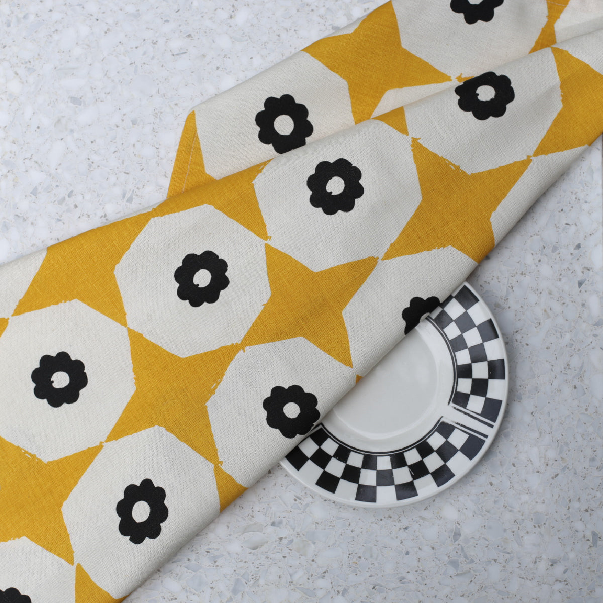 Closeup of yellow star and black flower printed hemp tea towel on a marble benchtop with black & white plate.
