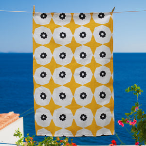 A yellow star and black flower printed hemp tea towel hanging on a clothesline with the deep blue sea behind and red flowers in the background