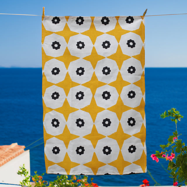 A yellow star and black flower printed hemp tea towel hanging on a clothesline with the deep blue sea behind and red flowers in the background