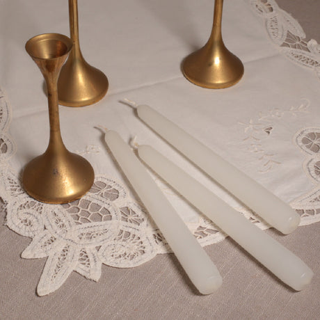 A set of three white Venetian-style tapered candles with gold-colored candleholders displayed on a lace tablecloth.