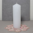 A white pillar candle on a pink lace doily with a grey background.