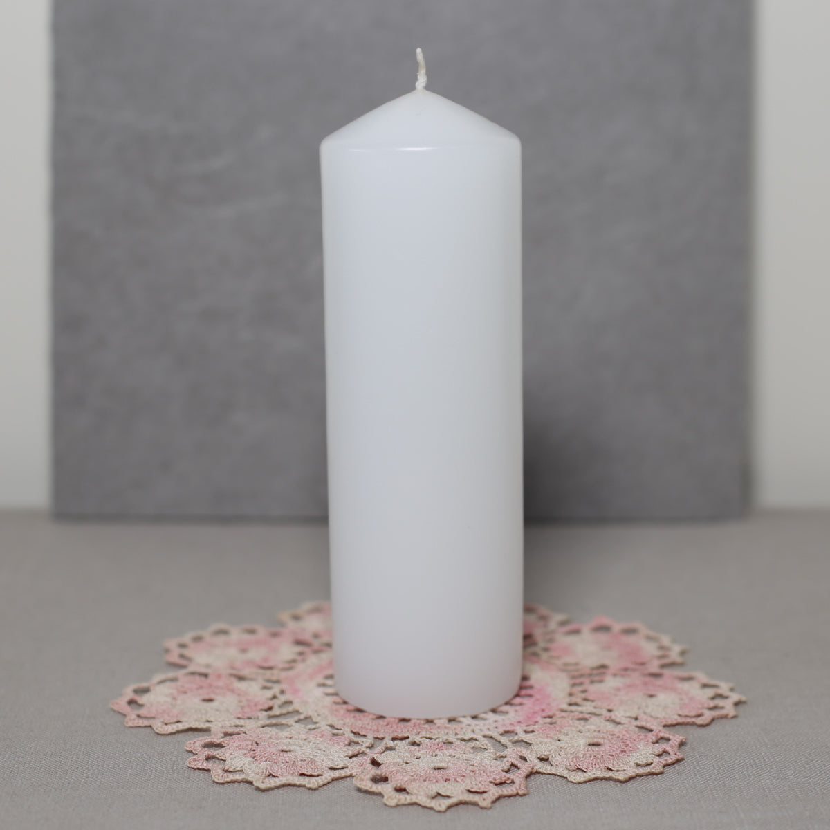 A white pillar candle on a pink lace doily with a grey background.