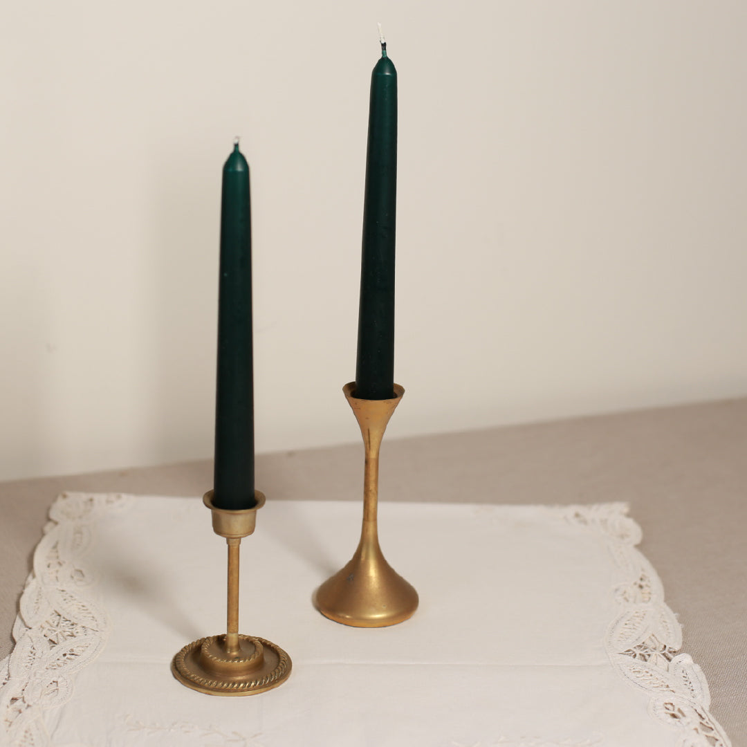 Two tapered Venetian-style table candles with green wax and gold-colored metal sticks, displayed on a lace tablecloth.