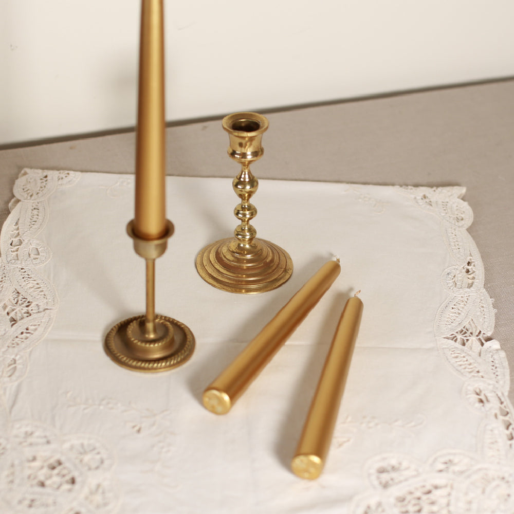 A gold-colored tapered Venetian-style table candle with metallic finish, accompanied by a candle holder and multiple wicks, displayed on a lace tablecloth.
