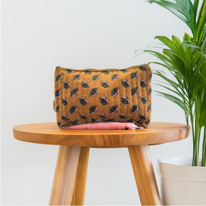 A large cosmetic pouch with a floral pattern in mustard, baby pink, and dark olive green, displayed on a wooden stool with a pink blanket on top, placed against a white wall and beside a green potted plant.