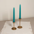 Two tapered Venetian-style table candles with turquoise wicks and bronze metal bases, placed on a lace-covered surface.